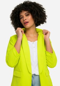 Blazer jaune-vert vif avec col à revers, deux poches avant et une texture lisse, porté sur une chemise blanche et un jean clair.