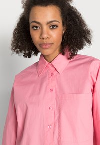 Camisa cor-de-rosa abotoada com colarinho pontiagudo, um único bolso no peito e mangas compridas. O tecido parece liso, com um leve brilho.
