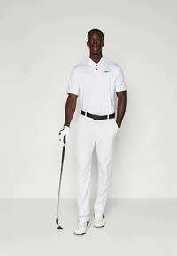 Λευκή γκολφ ενδυμασία που περιλαμβάνει ένα πόλο μπλουζάκι με γιακά και δύο κουμπιά, σε συνδυασμό με εφαρμοστά παντελόνια. Ο γκολφάς κρατάει ένα μπαστούνι και φοράει γκολφ παπούτσια.