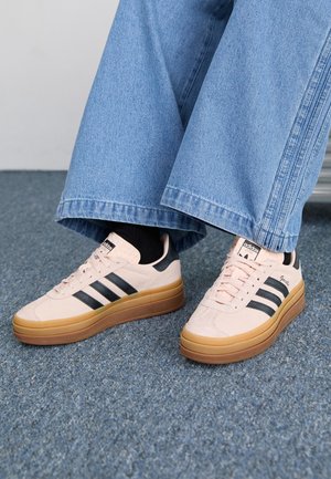 Pieds portant des baskets Adidas Gazelle beige avec des bandes noires et des semelles en gomme, associées à un jean bleu à jambes larges et des chaussettes noires sur un tapis.