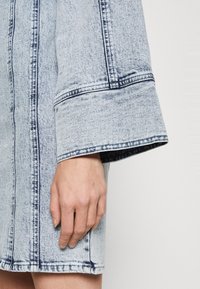 Světle modré džíny šaty s rozšířeným rukávem, se zvýrazněným šitím a texturovanou látkou. Ruka se dotýká látky poblíž lemu.