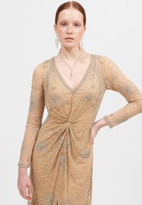 Beige, lange mouwen jurk met ingewikkelde zilveren kralen, een V-halslijn en een gedraaide ontwerp bij de taille, met een elegante vloeiende textuur.