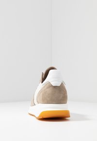 Beige ja valkoinen nubukivinen tennari, jossa on valkoinen kantanauha, johon on merkitty "Garment Project" ja oranssi pohja, kuvattuna takaa valkoisella pinnalla.