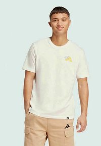 Λευκό βαμβακερό T-shirt με κοντά μανίκια, στρογγυλή λαιμόκοψη, περιέχει κίτρινο σχέδιο στην αριστερή θηλή και μαύρη ετικέτα λογότυπου κοντά στο στρίφωμα.