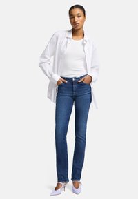 Chemise blanche boutonnée sur un débardeur blanc, jeans bleus ajustés et chaussures à talons hauts violet clair. La tenue présente un style décontracté.