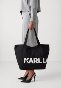 Czarna torba na ramię z płótna z białym logo "KARL LAGERFELD", wyposażona w dwa długie uchwyty i przestronny design, ułatwiający noszenie.