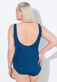 Un maillot de bain une pièce bleu profond avec de larges bretelles, un dos plongeant et un motif texturé sur le tissu.