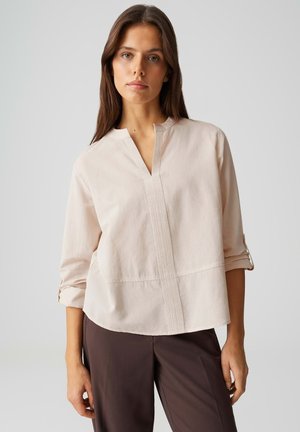 Donna con capelli castani lunghi che indossa una blusa beige chiaro a maniche lunghe e pantaloni marrone scuro, in piedi contro uno sfondo chiaro e uniforme.