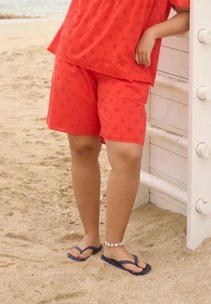 Osoba w czerwonych szortach z dziurkami i topie, granatowych klapkach oraz białej bransoletce z muszelkami na kostce, stojąca na piaszczystej plaży obok białej drewnianej konstrukcji.