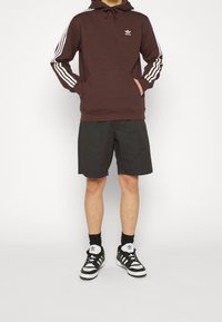 Sudadera marrón con rayas y logo blancos, combinada con pantalones cortos negros y zapatos en blanco y negro. La sudadera tiene un bolsillo canguro.