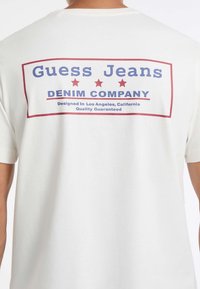 Osoba ubrana w białą koszulkę z czerwoną i niebieską grafiką oraz napisem "Guess Jeans Denim Company" na plecach.