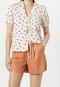 Blusa corta de manga corta en crema con patrones de rosas rojas y cierre con lazo. Combinada con pantalones cortos naranjas. Tela texturizada con un ajuste relajado.