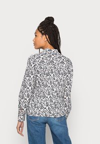 Camisa de botão com padrão floral, com uma base branca, flores azul-marinho e pequenos detalhes coloridos. Apresenta mangas longas e um colarinho clássico.