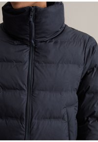 Veste matelassée bleu marine avec un col haut, fermeture éclair et texture matelassée. Comprend des poches latérales et une couleur unie sans motifs.