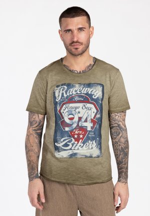Man met kort haar en tatoeages op de armen draagt een olijfgroen T-shirt met een vintage "Raceway 94 Bikers" afbeelding en beige broek.