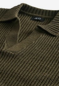 Olivovo zelený rebrovaný úplet s výstrihom do V a prehnutým golierom. Textúrovaná tkanina s viditeľnými švovými detailmi. Viditeľná značka.