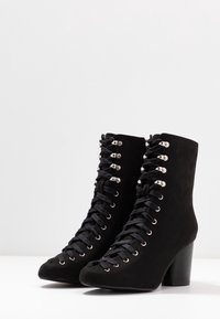 Botines de ante negro con puntera afilada, que presentan ojales y cordones plateados. Tacón grueso de mediana altura para mayor soporte.