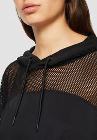 Gros plan d'une personne portant un sweat à capuche noir avec un panneau en mesh sur la poitrine et les épaules et des cordons pendants sur la capuche.