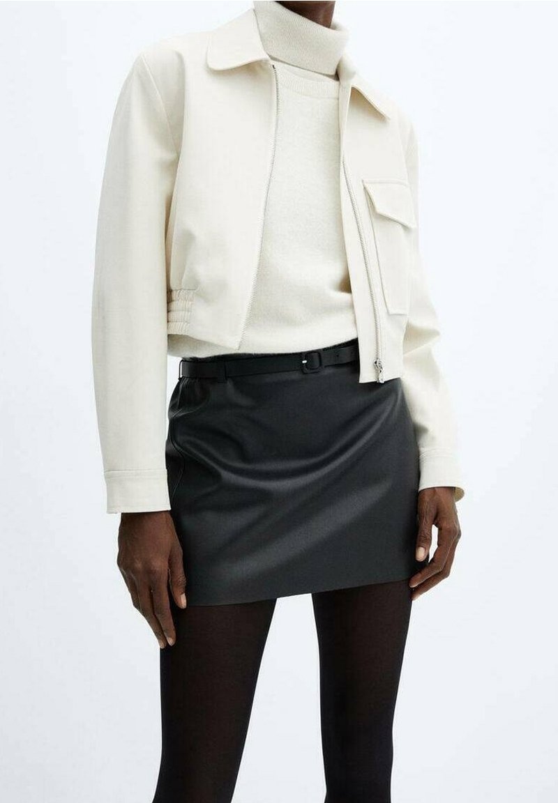 Personne portant une veste courte crème sur un col roulé crème, une ceinture noire, une mini-jupe noire et des collants noirs sur un fond blanc uni.