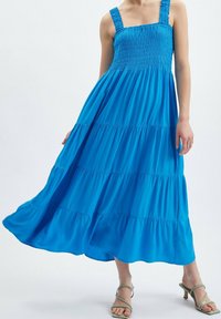 Blå, lång maxiklänning med smockad byst och tjocka, texturerade axelband. Har lager av volanger och en svängande silhuett.