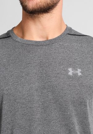 Mann trägt graues Under Armour T-Shirt mit sichtbarem Logo auf der Brust und teilweise Bart am Hals und Kinn.