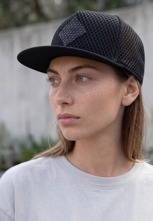 Mlada žena s blagim pjegama, nosi crnu mrežastu kapu i bijelu majicu, gleda u stranu na vanjskoj, zamagljenoj pozadini.