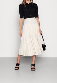 Plisserad krämfärgad kjol med svart kortärmad skjorta med knappar. Bärs med svarta sandaler med remmar och håller en liten svart handväska.