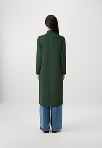 Cardigan lungo a coste verdi con colletto alto, maniche aderenti e spacchi laterali, abbinato a jeans in denim a gamba larga di colore blu chiaro.