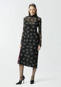 Crna mrežasta haljina dugih rukava, spiralan uzorak, visoki izrez, dužine do pola lista, s kontrastnim ružičastim bočnim panelom. Obuvene crne štikle.