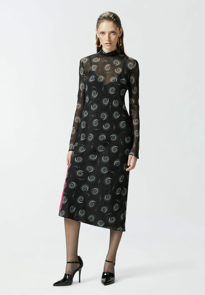 Crna mrežasta haljina dugih rukava, spiralan uzorak, visoki izrez, dužine do pola lista, s kontrastnim ružičastim bočnim panelom. Obuvene crne štikle.