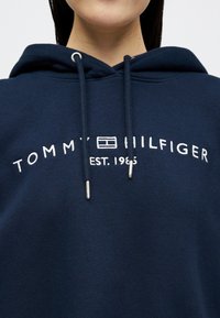 Σκούρο μπλε φούτερ με λευκή κεντημένη επιγραφή "Tommy Hilfiger EST. 1985" και λογότυπο, φορεμένο από ένα άτομο που εμφανίζεται από το πηγούνι μέχρι το στήθος.