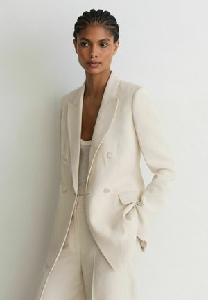 Vrouw met gevlochten haar die een crèmekleurige dubbelgeknoopte blazer en bijpassende broek draagt, staand tegen een witte muur met één hand in de zak.