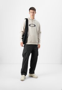 Mandlig model står i en minimalistisch miljø, iført en beige hoodie med et grafisk logo, sorte cargo-bukser og cremede sneakers.