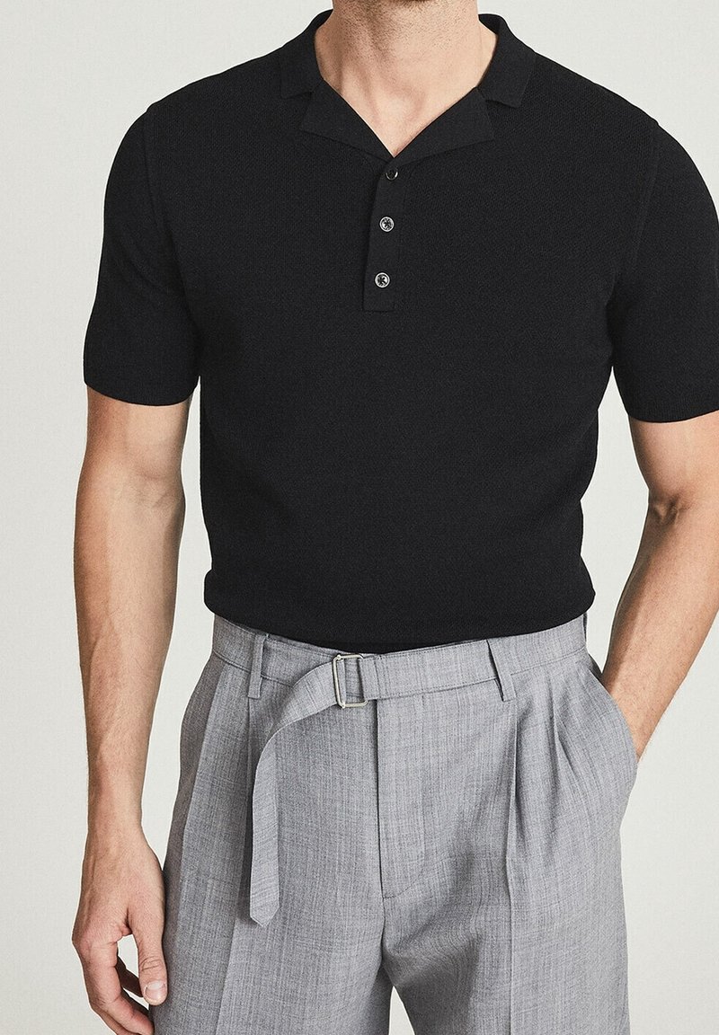Černé pletené polo tričko s krátkým rukávem a tříknoflíkovou légou, v kombinaci se světle šedými skládacími kalhotami a páskem.