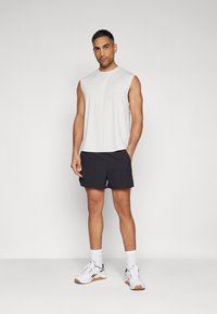 Hellgraues ärmelloses Oberteil, marineblaue Shorts, weiße Sneaker mit schwarzen Akzenten und knöchellange weiße Socken. Das Model steht vor einem neutralen Hintergrund.