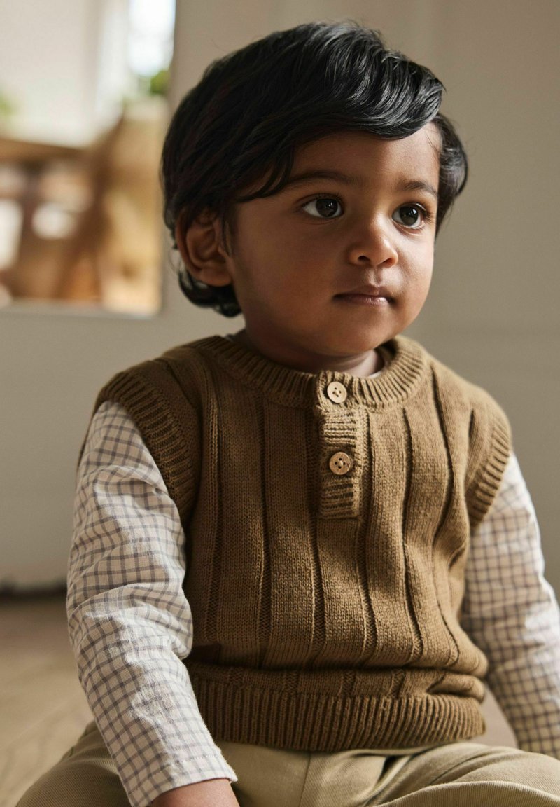 Jeune enfant aux cheveux foncés portant un gilet marron en tricot par-dessus une chemise à carreaux à manches longues, assis à l'intérieur avec une expression neutre.