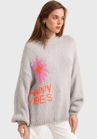 Pull en tricot gris à manches longues, avec un motif de fleur rose et "HAPPY VIBES" en orange, col et poignets côtelés.