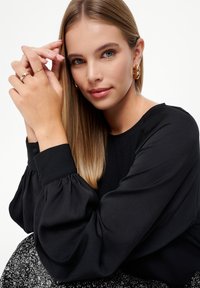 Čierna blúzka s dlhými rukávmi, vyp puffie rukávy, okrúhly výstrih a zapínanie na gombíky na zápästiach. Model nosí zlaté kruhové náušnice a vrstvené prstene.