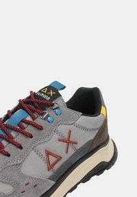Sneaker in suede e tessuto grigio con lacci rossi, accenti blu e logo geometrico. Presenta una suola testurizzata e occhielli rinforzati.