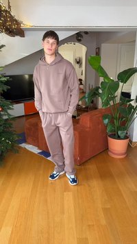 Καφέ φούτερ με κουκούλα και ταιριαστά παντελόνια φόρμας από απαλό ύφασμα, σε συνδυασμό με μπλε και λευκά αθλητικά παπούτσια. Εσωτερικός χώρος με πράσινο φυτό και έπιπλα.