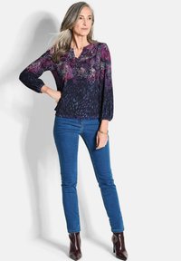 Langärmlige Bluse in Dunkelblau mit pinken und lila Blumenmustern, kombiniert mit engen blauen Jeans und bordeauxfarbenen Stiefeletten mit Absatz.