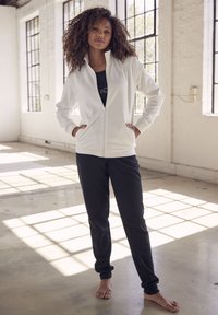 Weiße Zip-Jacke mit hohem Kragen, kombiniert mit schwarzen Hosen. Der Stoff ist glatt, hat seitliche Taschen und gerippte Bündchen.