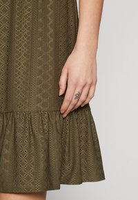 Oliivinvihreä mekko, jossa on teksturoitu, silmäkuvioitu kangas, liehukehelmakanttaminen helma ja vartalonmyötäinen yläosa. Käsi esillä, jossa pieni tatuointi.