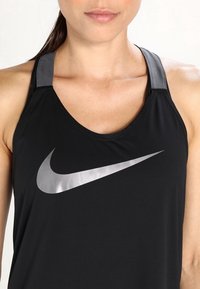 Nainen, joka on pukeutunut mustaan Nike-hihattomaan paitaan, jossa on hopeinen swoosh-logo, näyttäen kaula-aluetta, hartioita ja osan kasvoista suljetuilla huulilla.