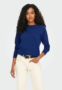 Kuningakindel sinine kudum, millel on ümar kaelus ja pikad varrukad, koos hallide beežide pükste ja musta vöödiga. Kujundatud istuvaks välimuseks.