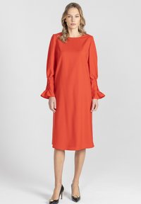 Czerwona sukienka do kolan wykonana z gładkiej tkaniny, z d�ługimi rękawami ze zdobieniami marszczącymi przy mankietach i zaokrąglonym dekoltem. Założone czarne szpilki.