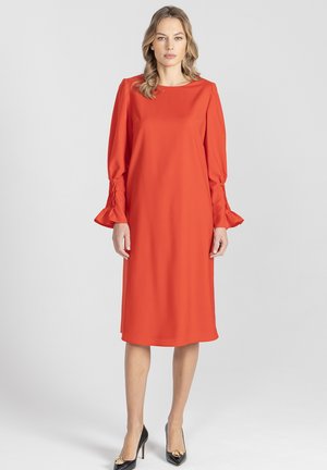 Czerwona sukienka do kolan wykonana z gładkiej tkaniny, z długimi rękawami ze zdobieniami marszczącymi przy mankietach i zaokrąglonym dekoltem. Założone czarne szpilki.