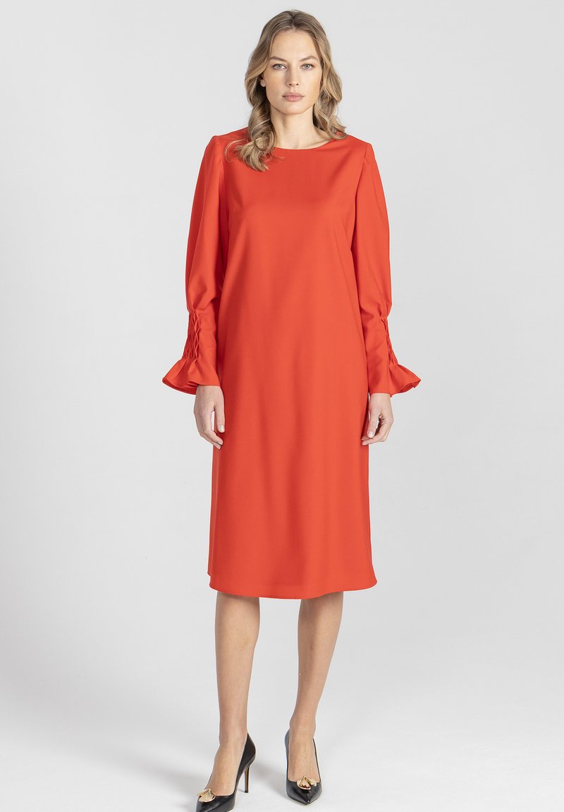 Czerwona sukienka do kolan wykonana z gładkiej tkaniny, z długimi rękawami ze zdobieniami marszczącymi przy mankietach i zaokrąglonym dekoltem. Założone czarne szpilki.