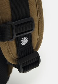 Brązowy worek z tkaniny z czarnym paskiem z taśmy, ozdobiony plastikową klamrą i wyraźnym, białym haftowanym logo na naszywce.