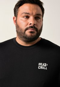Hombre con barba vestido con una camiseta negra que dice "RELAX + CHILL", mirando ligeramente de lado contra un fondo liso claro.